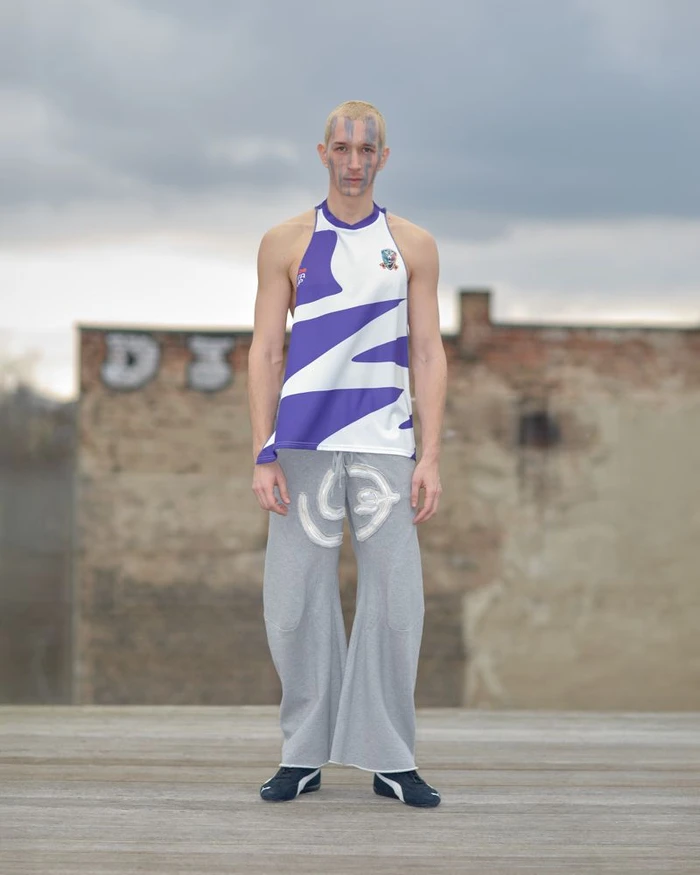 Model standing outdoors wearing a purple and white top and grey trousers against a brick wall and cloudy sky