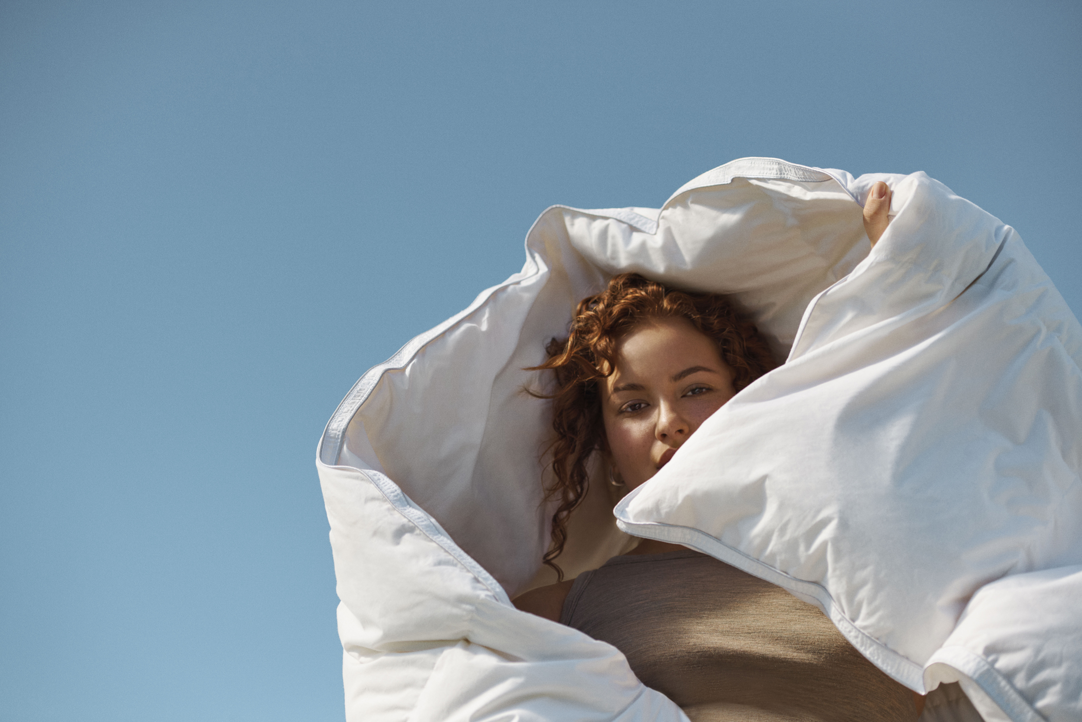 Woman with red curly hair is partially covered by a white duvet against a clear blue sky in a low-angle shot in an outdoor setting.