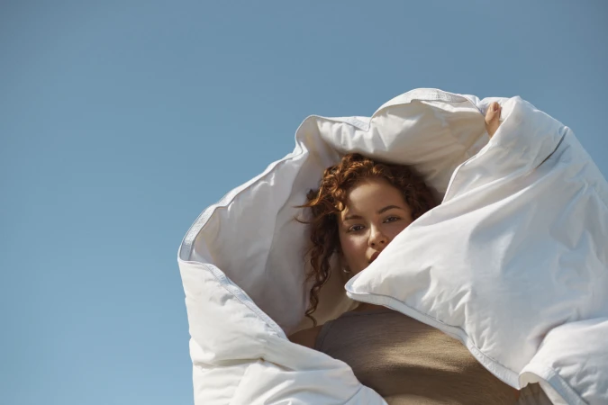Woman with red curly hair is partially covered by a white duvet against a clear blue sky in a low-angle shot in an outdoor setting.
