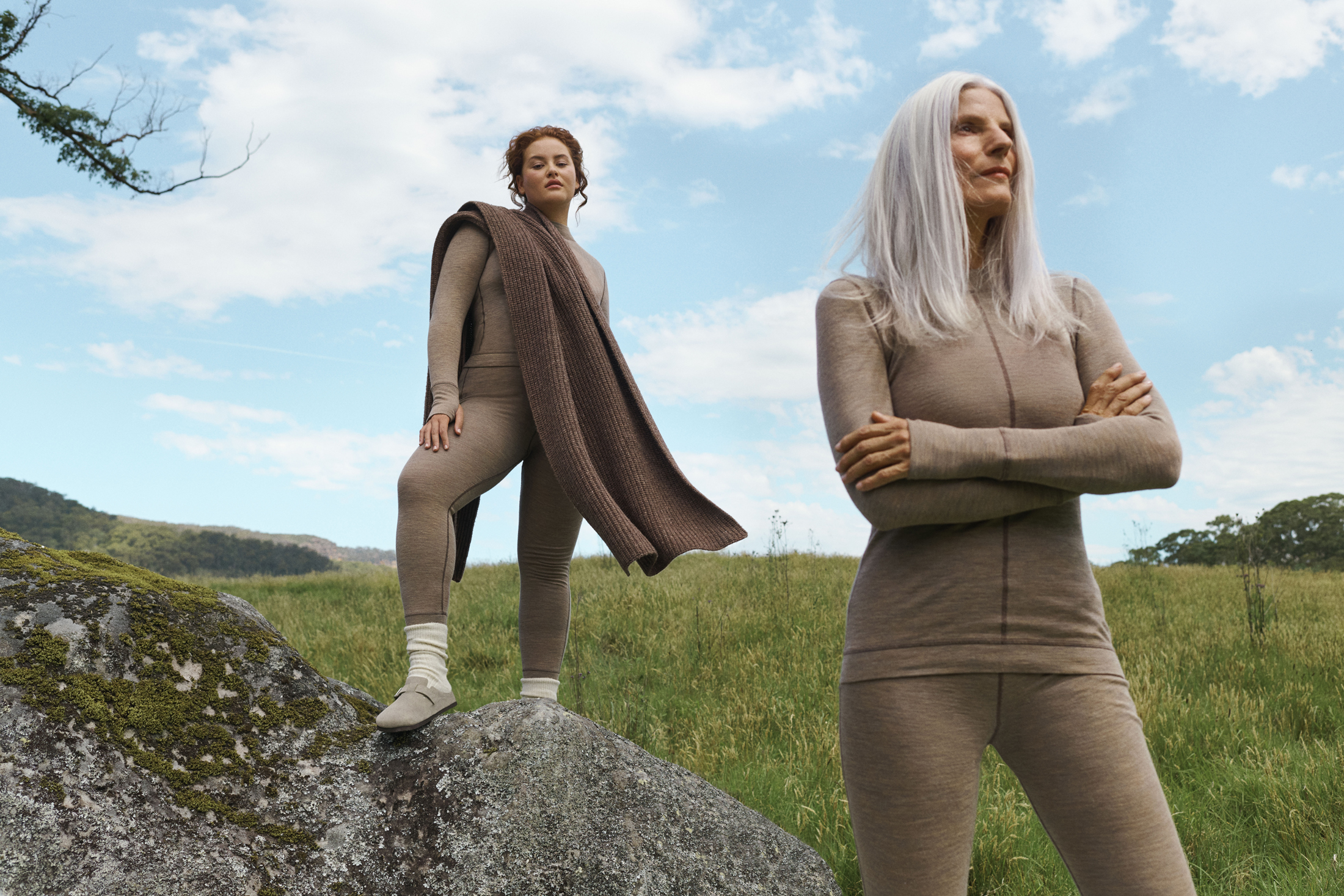 Two adult women in natural light wearing matching activewear pose in a green field, one standing on a mossy rock, and clear blue sky.