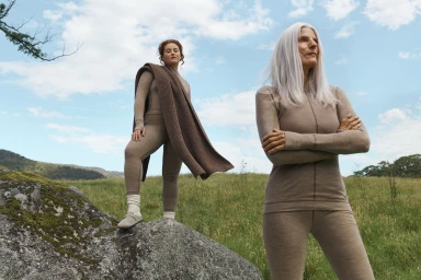 Two women wearing matching beige knitwear outdoors in a green meadow beneath a partly cloudy sky.