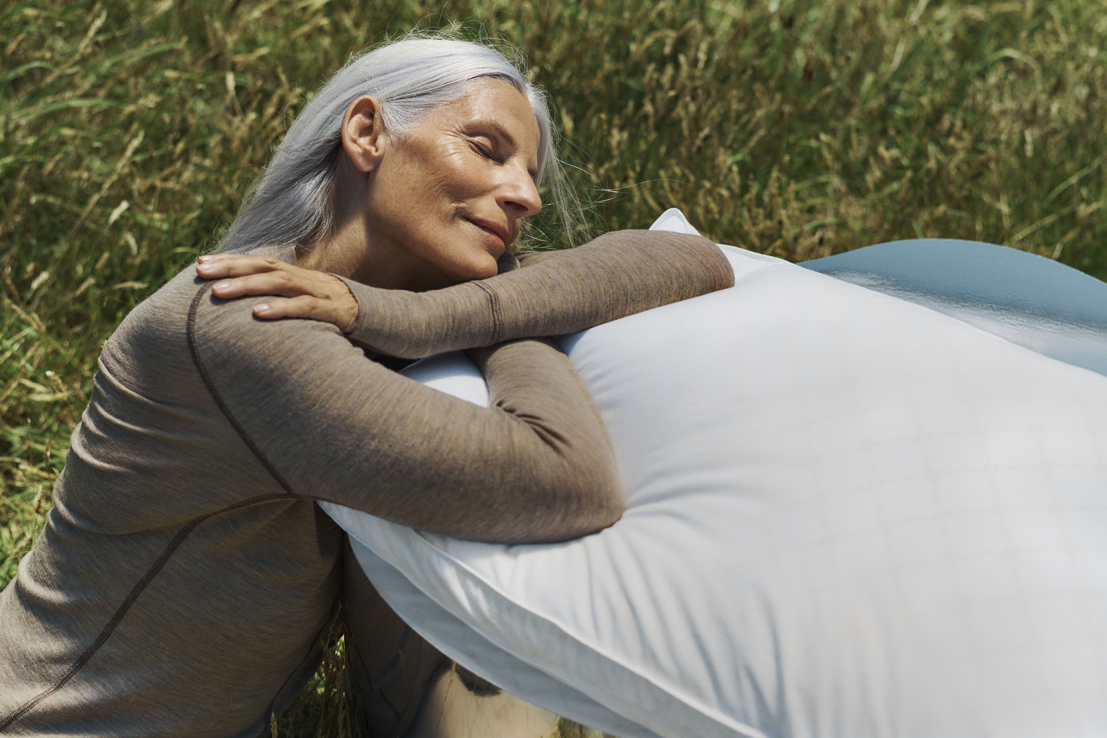 Adult woman with long gray hair reclining on a large white pillow outdoors surrounded by tall green grass in warm daylight.