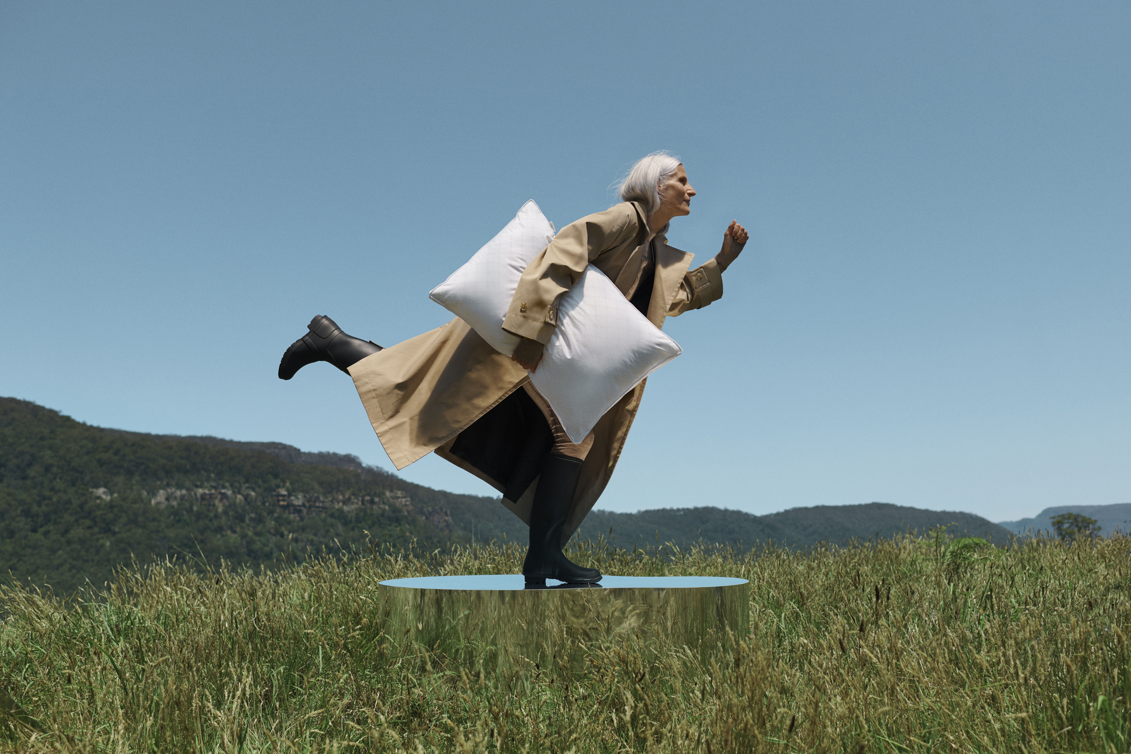 A mature woman in a trench coat balances on a reflective platform in a grassy field, holding two pillows against a blue sky.