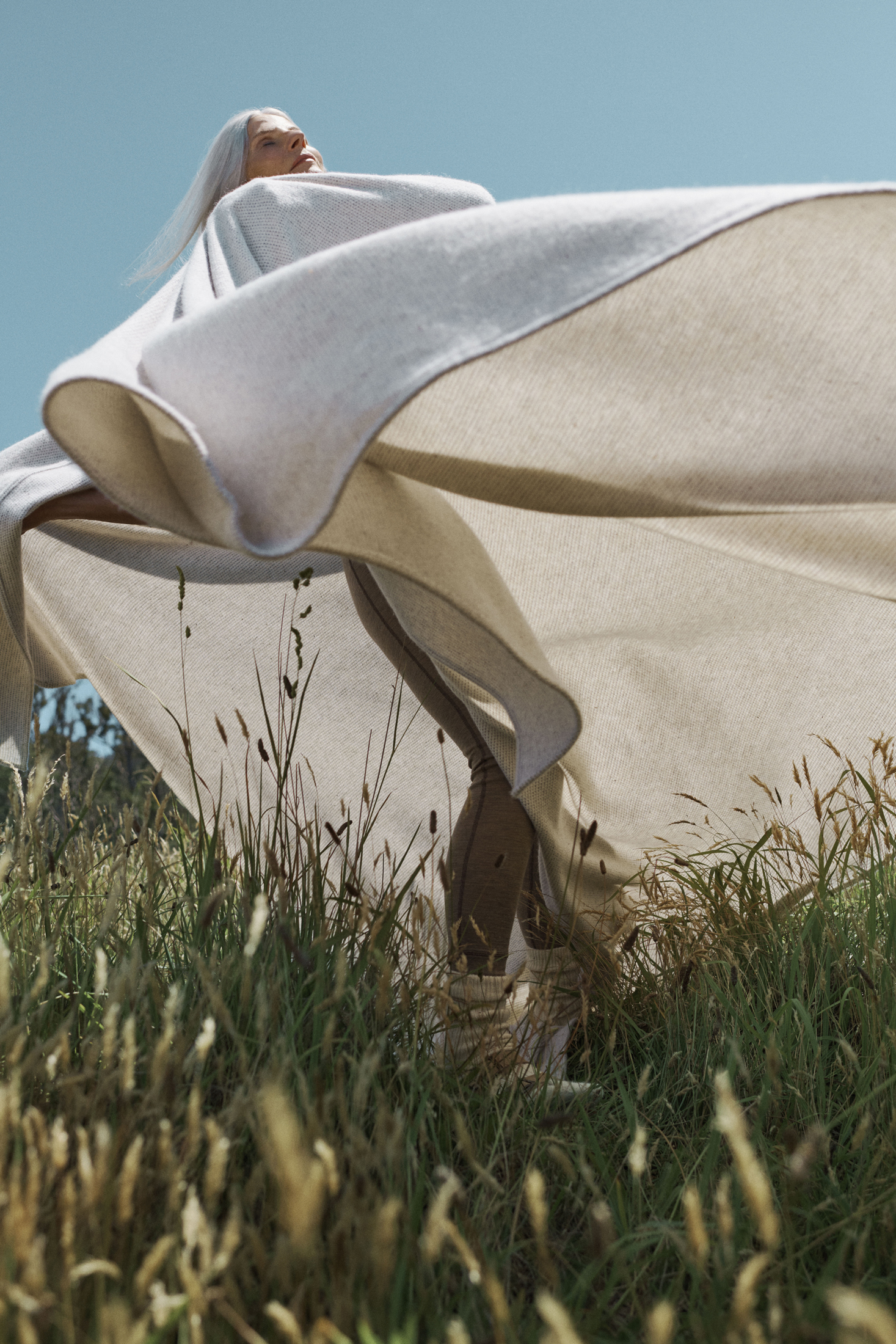 Woman with long gray hair poses outdoors in a field, wrapped in a beige shawl, beneath a blue sky, captured in a sunlit fashion shot.