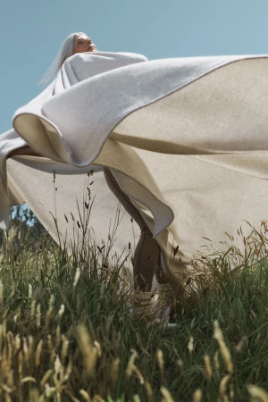 Woman with long gray hair poses outdoors in a field, wrapped in a beige shawl, beneath a blue sky, captured in a sunlit fashion shot.