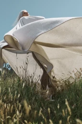 Older woman with long white hair wrapped in a large cream textile outdoors in a grassy field under a clear blue sky