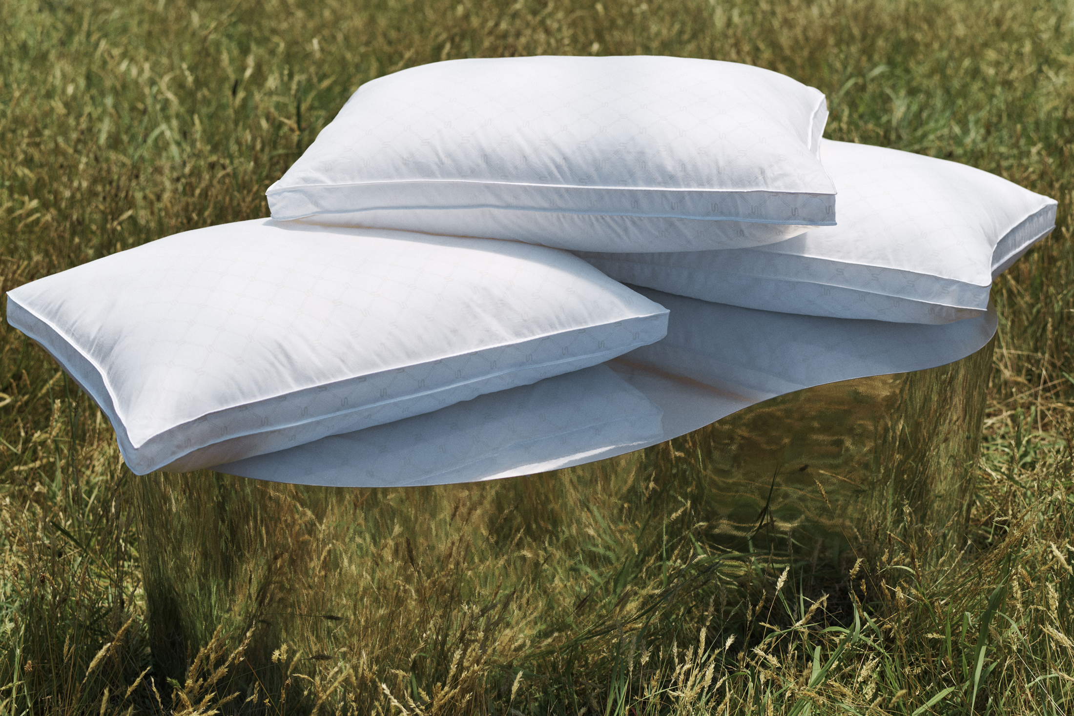 Three white pillows are stacked on a gold mirrored cylinder outdoors in a grassy field, creating a minimalist and luxurious feel.