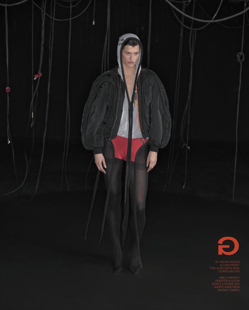 Fashion shot of a young adult man in an oversized black jacket, red underwear, and sheer black stockings, posing against a black backdrop.