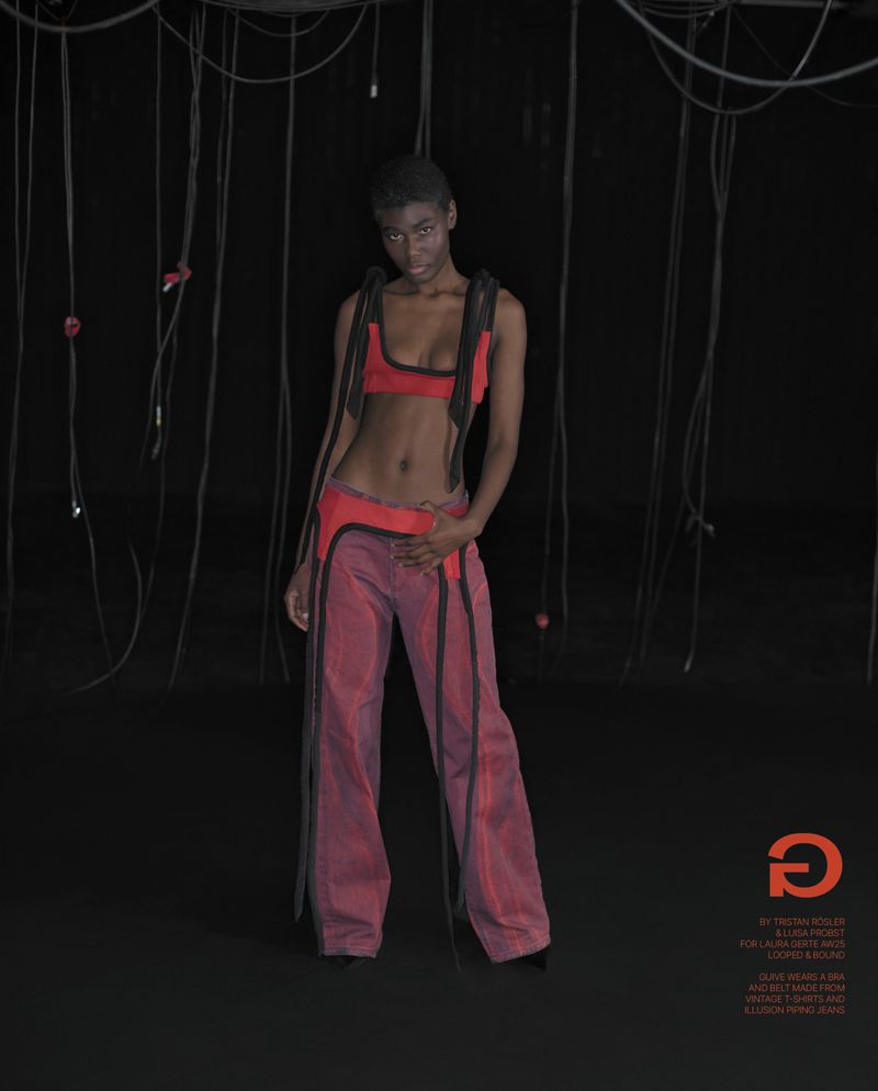 An adult woman poses indoors with wires in a red bra top and loose pants, looking at the camera against a dark background.