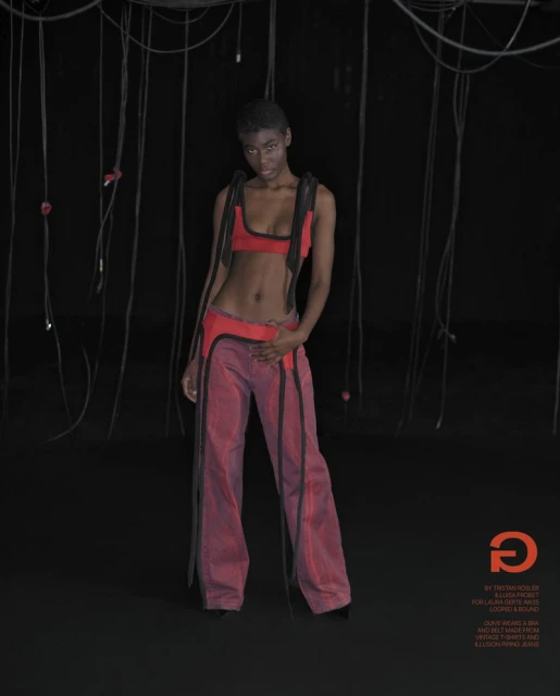 An adult woman poses indoors with wires in a red bra top and loose pants, looking at the camera against a dark background.