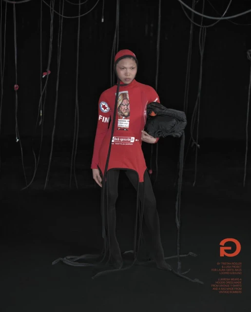 Fashion model in red long-sleeve shirt with attached leggings poses indoors with hanging cords and a black accessory bag.