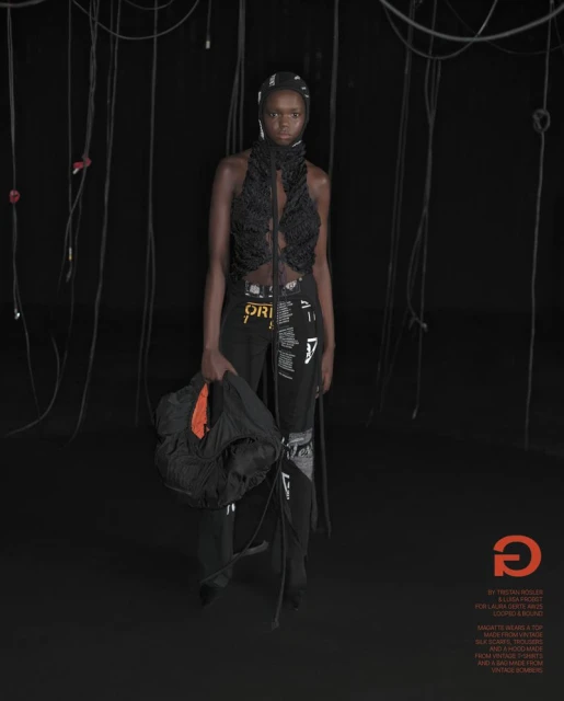 A dark-skinned woman poses in avant-garde fashion in a studio, wearing a black textured top and pants with graphic prints, indoor setting.