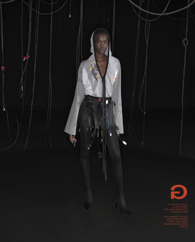 Fashion shot of a woman wearing a hooded top and a skirt with cables, standing in a dark studio with hanging cables.