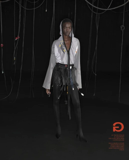 Fashion shot of a woman wearing a hooded top and a skirt with cables, standing in a dark studio with hanging cables.