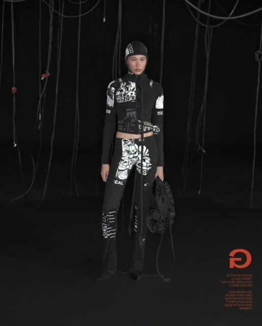 Fashion photo of a woman posing in a black printed jacket and pants ensemble with a matching hat, set against a dark background.