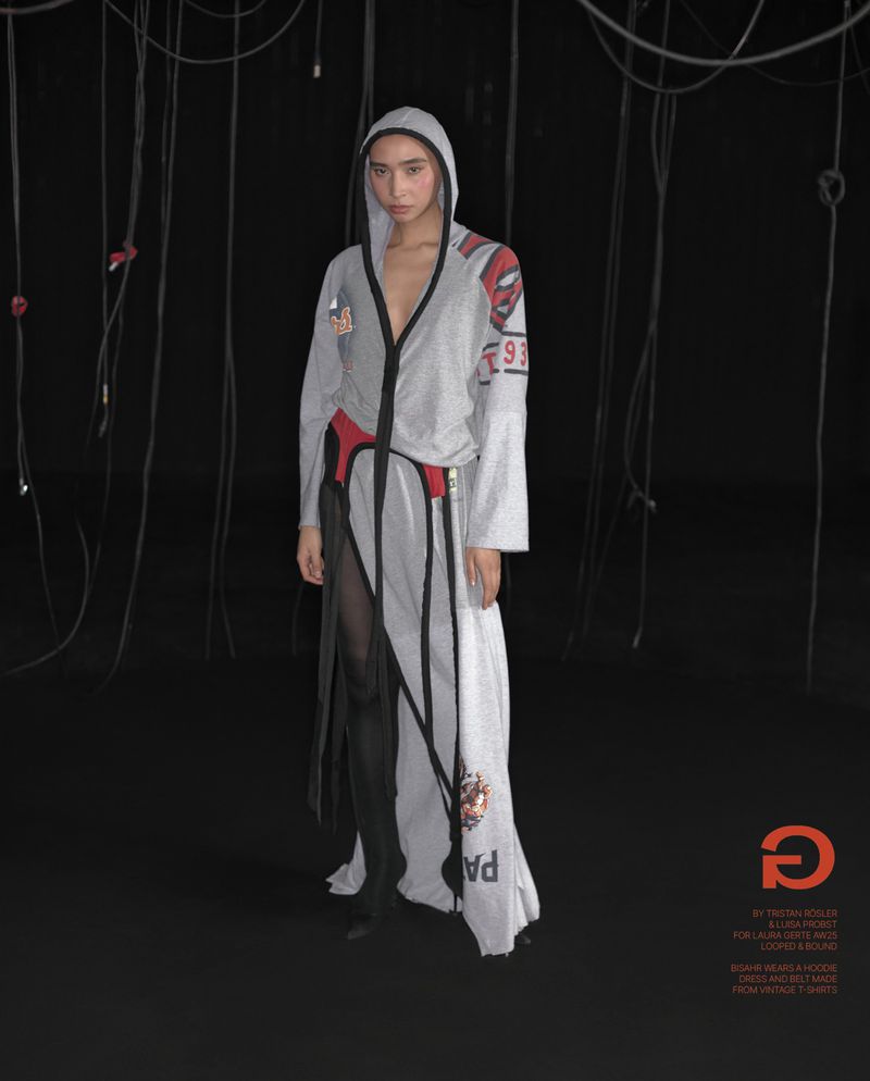 Fashion shot of a slender model posing in a long, grey hooded dress with black accents in front of a dark backdrop with hanging cords.