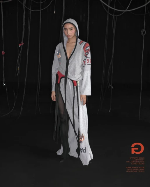 Fashion shot of a slender model posing in a long, grey hooded dress with black accents in front of a dark backdrop with hanging cords.