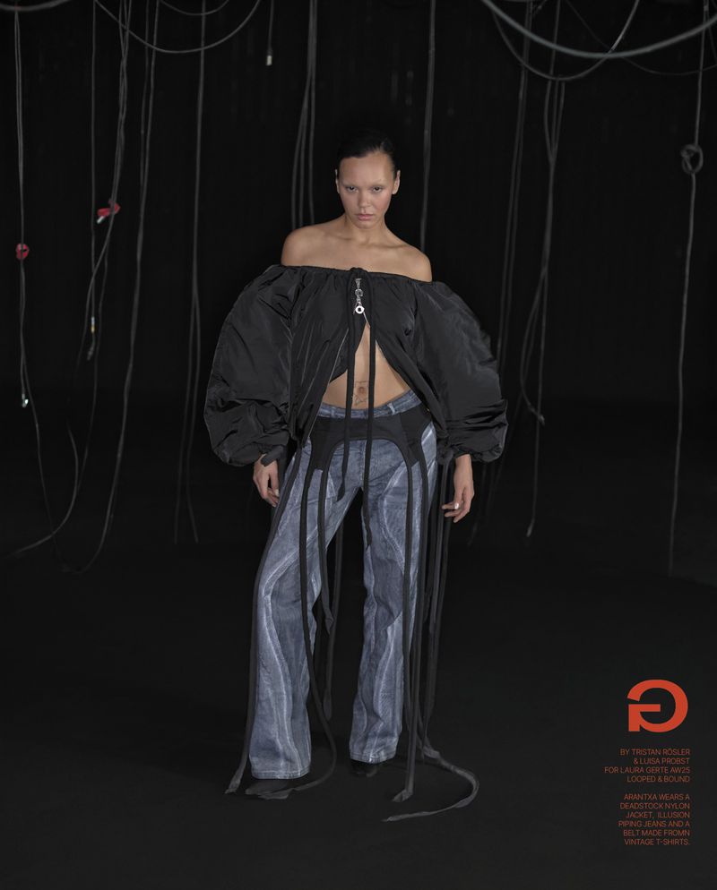 Fashion-forward studio shot: A lone adult female in a black, off-shoulder jacket and patterned pants stands against a dark backdrop.