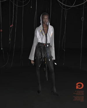 Model wearing a grey hooded top and cable-fringed skirt against a black backdrop with hanging cords.