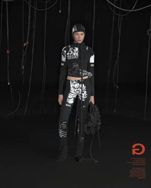 Model in black and white printed zip-up hoodie, trousers, and matching hood holding a black bag against a dark studio background