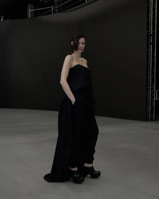 Adult female in an all-black strapless top and wide leg pants poses in a photo studio with a black backdrop and light grey floor.