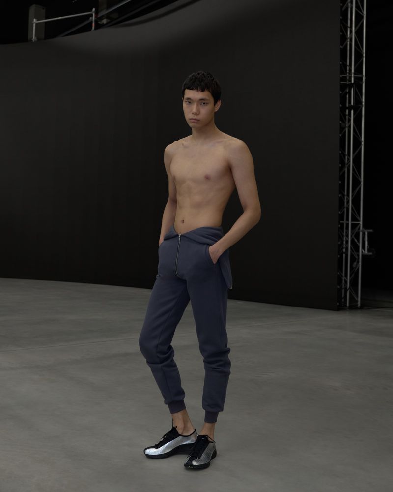 Man poses shirtless in grey sweatpants and athletic shoes against black backdrop in studio setting, full body shot, neutral expression.