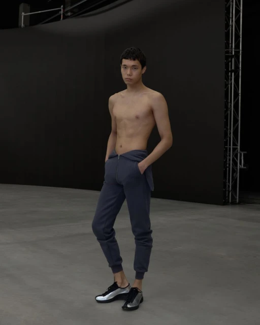 Man poses shirtless in grey sweatpants and athletic shoes against black backdrop in studio setting, full body shot, neutral expression.
