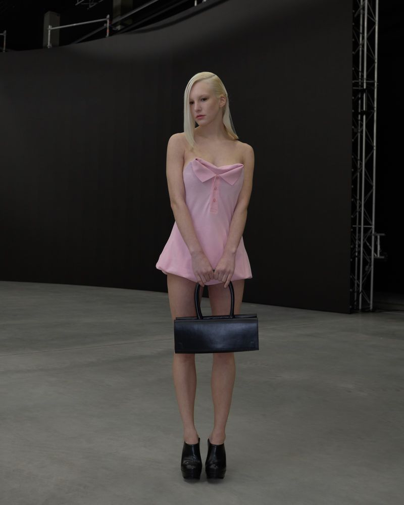 Full shot of a fair-skinned, adult woman with blond hair in a pink dress and black platform shoes holding a black purse.