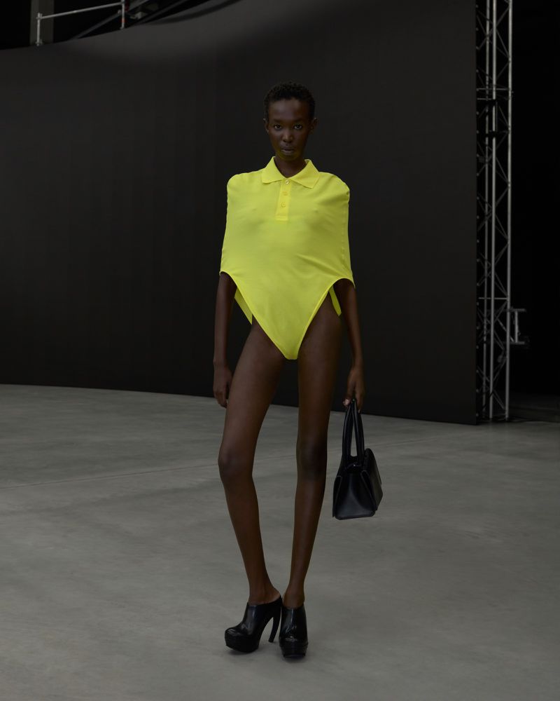 An adult woman models a yellow swimsuit with an attached polo shirt in a minimalist studio with a black background and concrete floor.