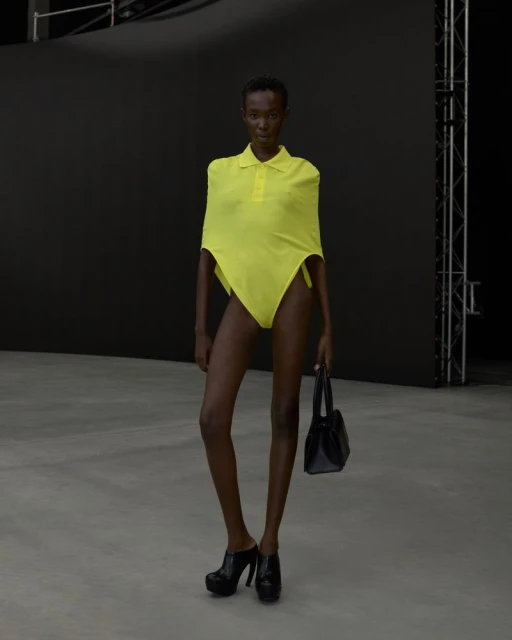 An adult woman models a yellow swimsuit with an attached polo shirt in a minimalist studio with a black background and concrete floor.