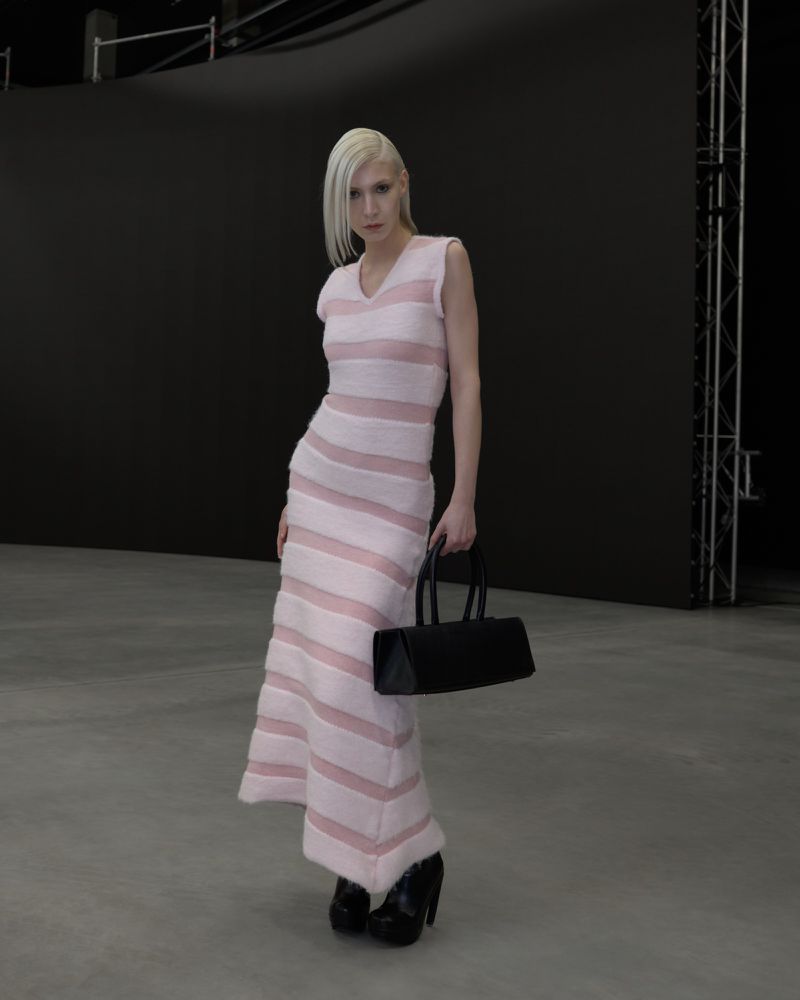 Model with blonde hair wearing a striped pink dress holding a black handbag in a studio space
