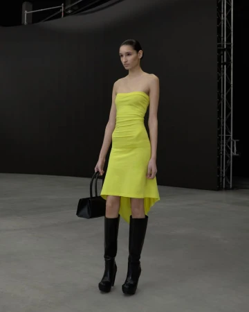 Model in strapless yellow dress holding a black handbag wearing tall black boots against a grey studio floor background