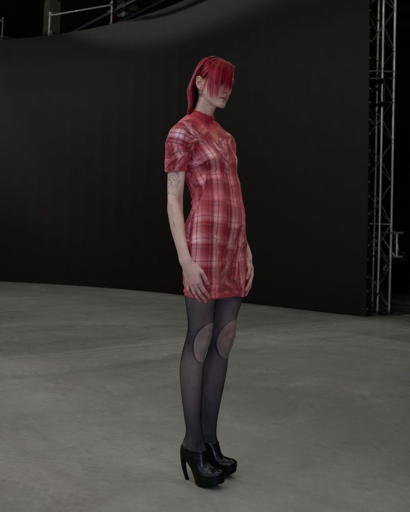 A slender woman with red hair poses in a short-sleeved plaid dress with black patterned stockings and high heels in a photo studio.