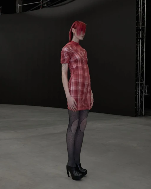 A slender woman with red hair poses in a short-sleeved plaid dress with black patterned stockings and high heels in a photo studio.