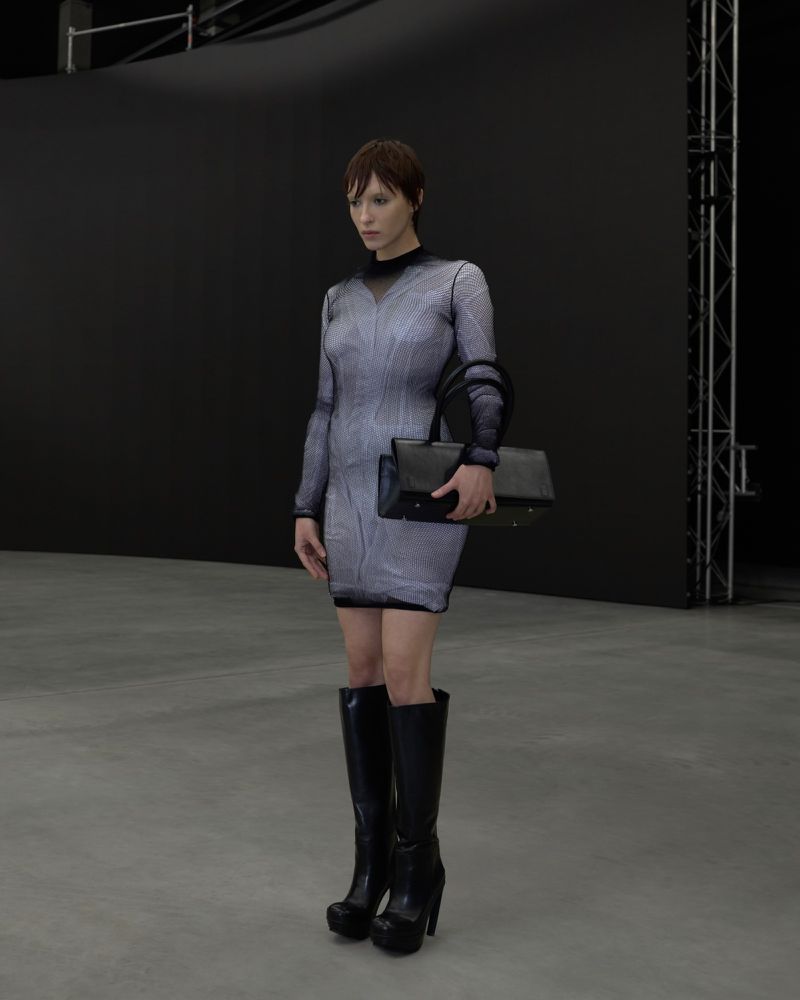 Model in a patterned mini dress and black boots holding a structured bag in a dark studio