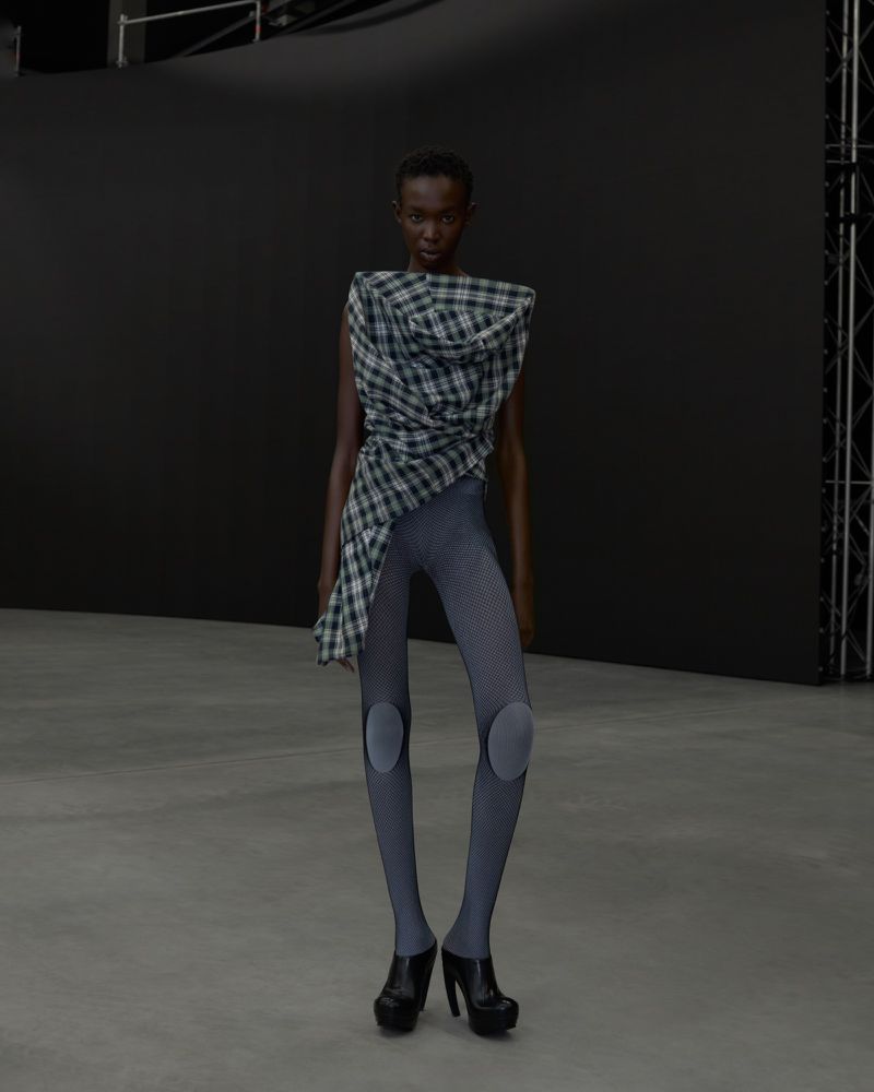 An African American woman models a draped plaid top, gray leggings with knee pads, and black platform heels against a dark background.