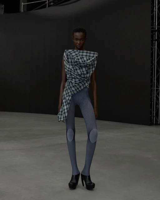 An African American woman models a draped plaid top, gray leggings with knee pads, and black platform heels against a dark background.
