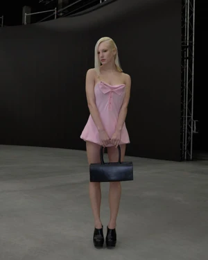 Model with blonde hair wearing a pink strapless mini dress and holding a black handbag in a studio
