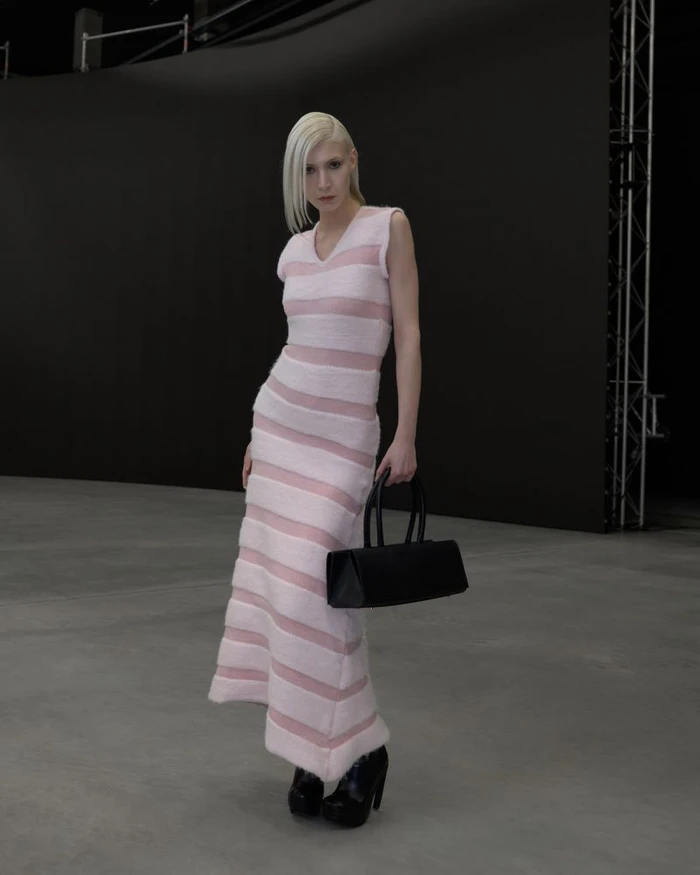 Model with blonde hair wearing a striped pink dress holding a black handbag in a studio space