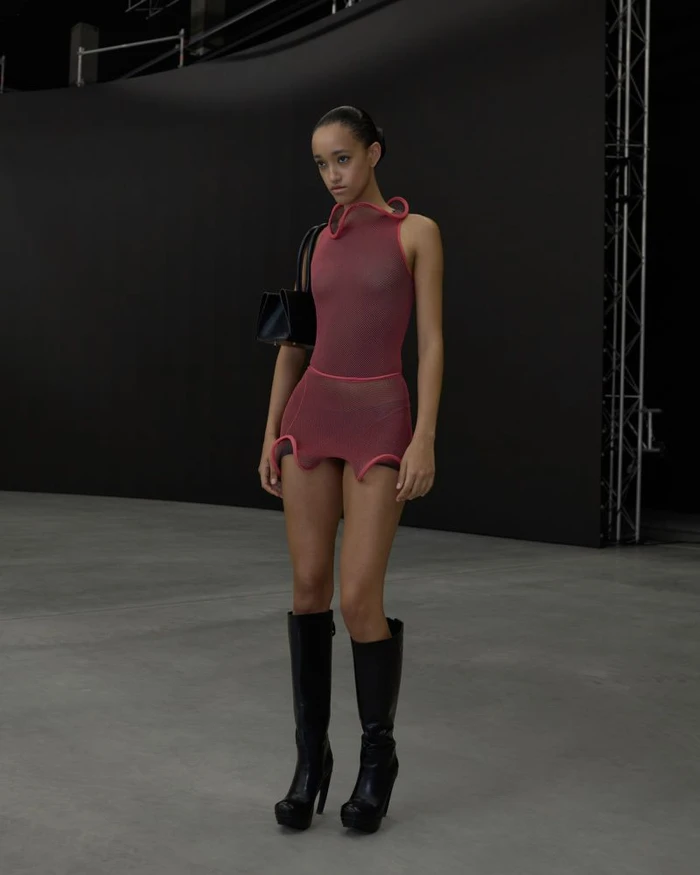 Model wearing a sheer pink sleeveless dress and black boots, carrying a handbag against a dark backdrop.
