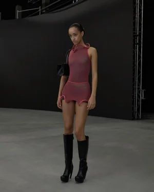 Model wearing a sheer pink sleeveless dress and black boots, carrying a handbag against a dark backdrop.