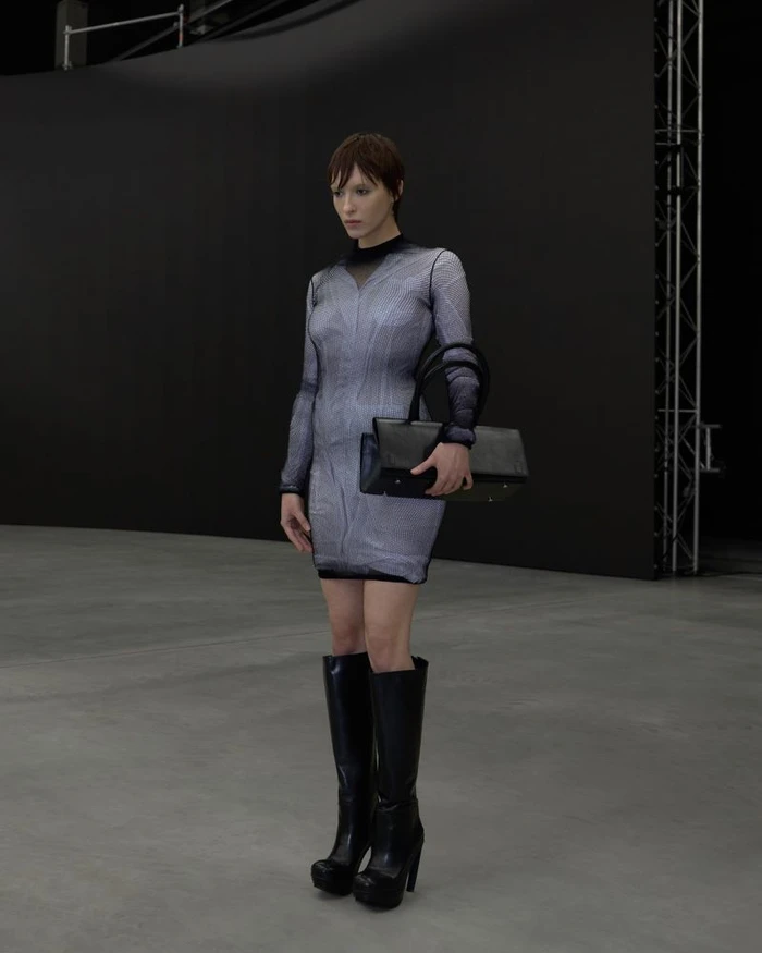 Model in a patterned mini dress and black boots holding a structured bag in a dark studio