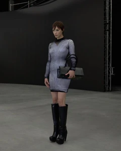 Model in a patterned mini dress and black boots holding a structured bag in a dark studio