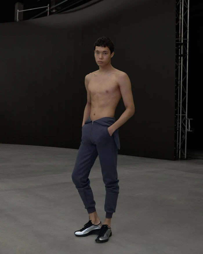 Shirtless male model wearing dark blue sweatpants and silver metallic sneakers standing in a studio.