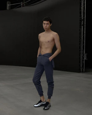 Shirtless male model wearing dark blue sweatpants and silver metallic sneakers standing in a studio.