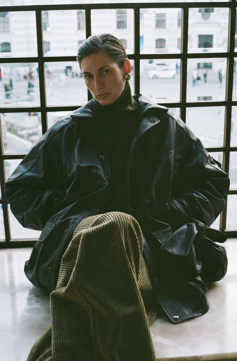 An adult woman is sitting by a window wearing a black turtleneck, a leather jacket and olive green corduroy pants.