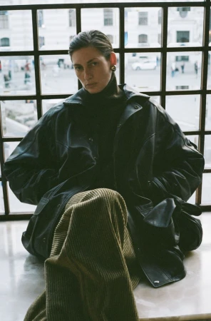 An adult woman is sitting by a window wearing a black turtleneck, a leather jacket and olive green corduroy pants.