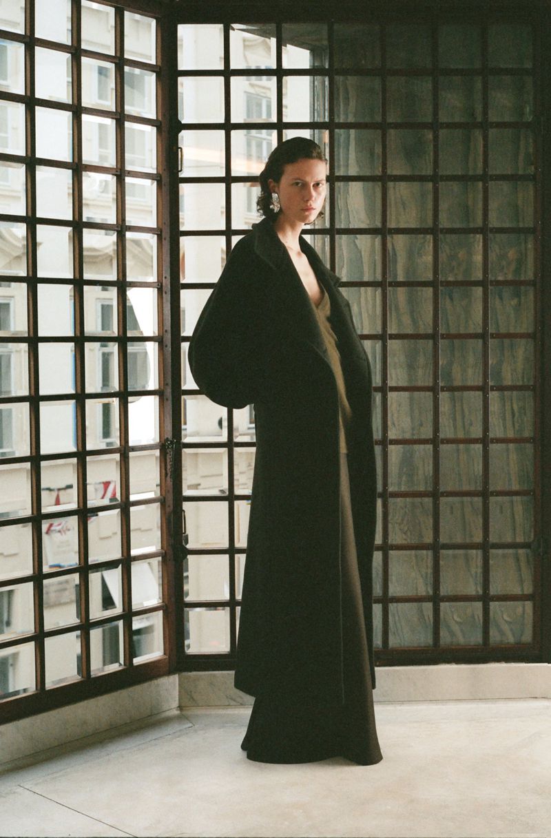 A woman stands in front of a grid-patterned window wearing a long black coat and a dress. She poses in an interior setting.
