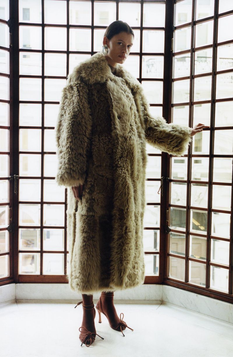 A stylish adult female poses indoors wearing a full-length, light-brown faux-fur coat and high-heeled shoes, standing near a window.