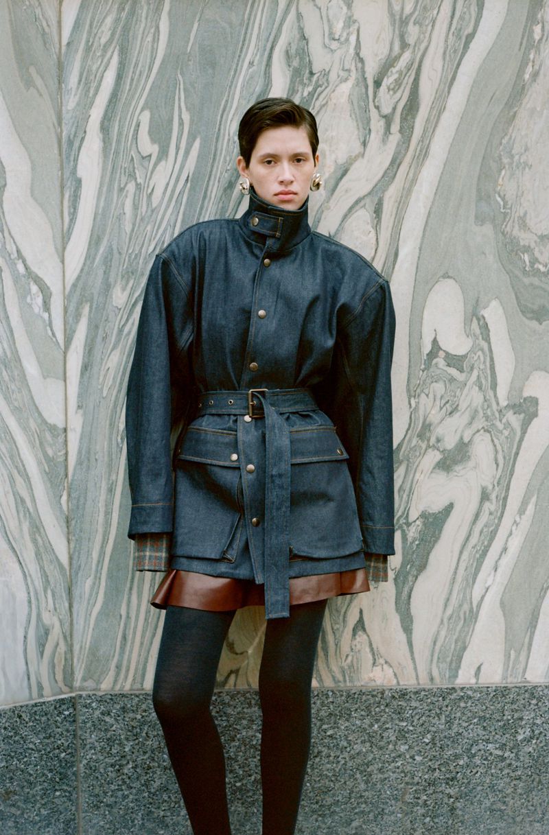 Model standing against patterned marble wall wearing a belted dark jacket and skirt